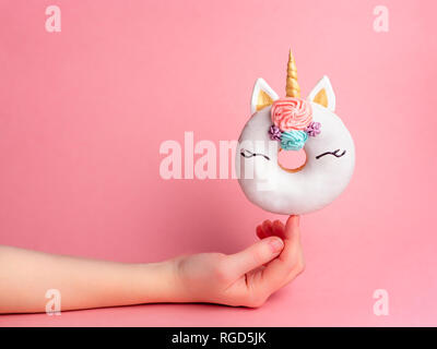 Unicorn donut on forefinger over pink background. Female hand hold trendy donut unicorn with white glaze on index finger. Copy space for text. Stock Photo
