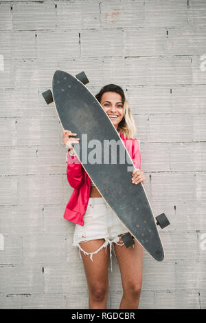 Portrait of a young woman with her long board, in front of a wall Stock Photo