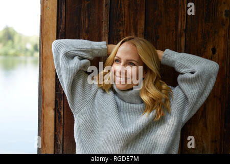 Smiling blond woman leaning against wooden hut at a lake Stock Photo