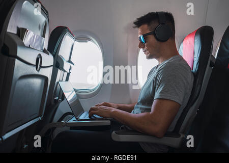 Man in airplane, using laptop, headphones Stock Photo