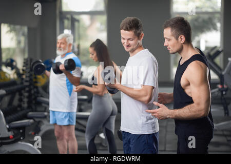 Muscular, athletic trainer helping man to do exercises with lifting dumbblells. Fit people wearing in sportswear. Female coach concentrated on elderly client working out. Stock Photo