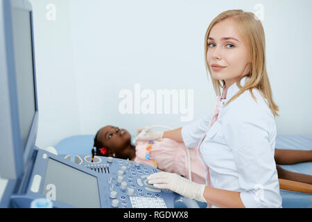 Portrait of two people. Little girl laying on bad during medial consultation in doctors office. Young professional doctor sitting and looking at camera during making diagnosis. Stock Photo