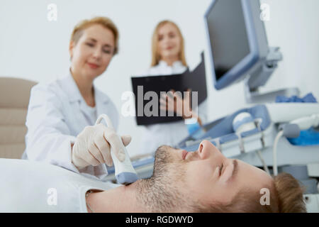 Closup crop view of three people in hospital. Young man on medical consultation. Adult professional doctor providing medical examination with spesial equipment and young nurse standing and helping. Stock Photo