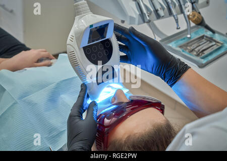 Procedure of teeth whitening in dentistry clinic. Woman wearing in protective glasses lying on dentist chair. Doctor holding modern device, ultraviolet lamp with blue light, Stock Photo