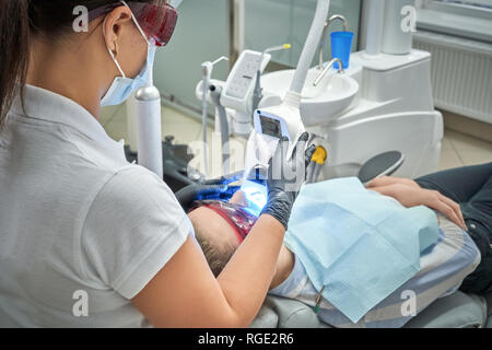 View from back of professional dentist in process of doing whitening of teeth to woman. Client lying on dental chair in protective glasses while doctor doing procedure with modern equipment. Stock Photo