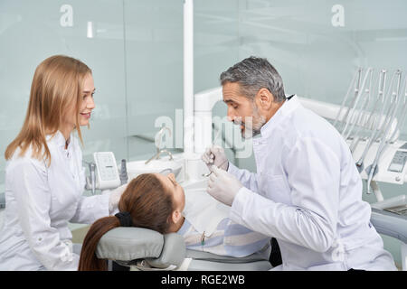 Stomatology doctor working with client in dentist cabinet. Young woman lying on dentist chair. Assistant helping, dentists wearing in white lab coats, holding special instruments. Stock Photo
