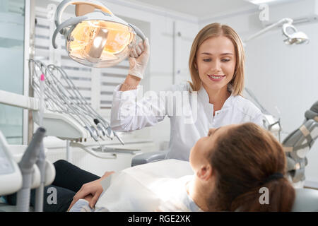 Beautiful woman working as dentist in clinc. Stomatologist holding hand on special lamp, looking at client. Patient of dentistry lying in dentist chair. Doctor wearing white coat and gloves Stock Photo
