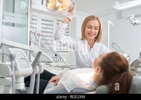 Patient lying on dentist chair and consulting with doctor. Beautiful woman in white uniform and gloves holding hand on special lamp, smiling, looking at client. Stock Photo