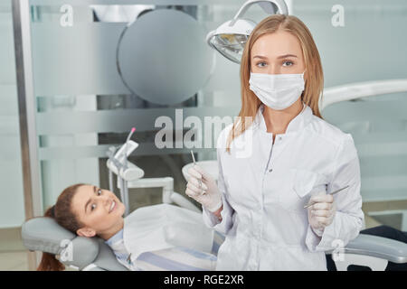 Professional doctor and happy patient posing, looking at camera. Dentist wearing in white coat, gloves and medical mask holding restoration instruments. Private dentistry clinic. Stock Photo