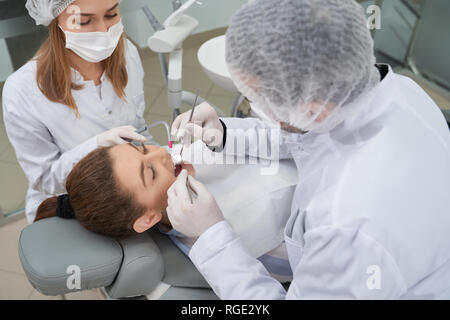 Patient lying with opened mouth on dentist chair in private clinic. Professional doctor preventing caries using special tools, restoration instruments. Assistant helping stomatologist. Stock Photo