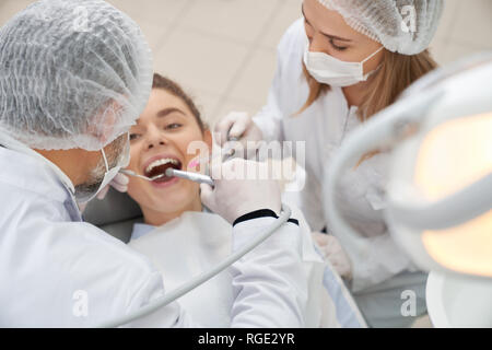 View from above of happy woman lying on dentist chair with opened mouth, Doctors wearing in white caps, masks and uniform working with client's teeth. Stomatologists using restoration tools.