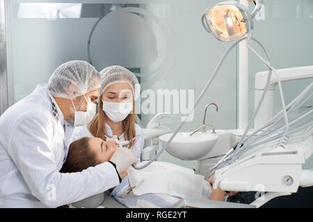 Modern stomatology private clinic with professional equipment. Dentists working with client's teeth. Woman lying on dentist cahir. Doctors wearing in medical caps, unforms and masks. Stock Photo