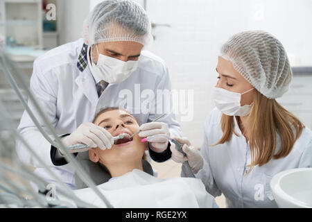 Dentist and assistant working with client. Woman lying with opened mouth on dentist chair. Stomatologists preventing caries using special restoration instruments and tools. Stock Photo