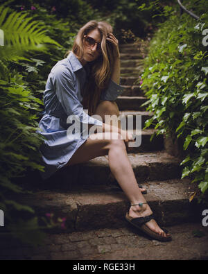 beautiful young girl in a blue dress sitting on stairs outdoors wearing sunglasses, short dress, legs showing Stock Photo