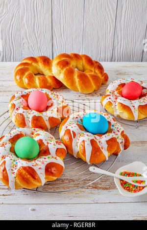 individual sweet Italian braided Easter Bread Rings glazed around dyed egg and topped with colorful sprinkles on a wire rack, on an old white wooden t Stock Photo