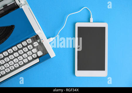 Flat lay of typewriter and blank screen digital tablet device connected with cable against blue background. Retro modern data transfer minimal creativ Stock Photo