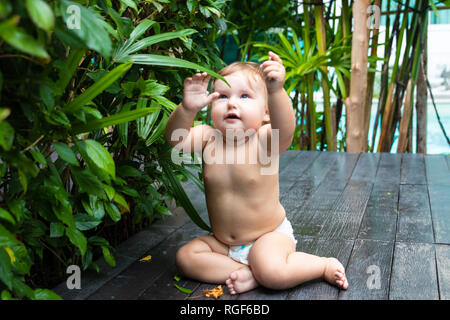 Pretty crawling baby girl smiling and crawling on the wooden brown walkway on the background of green bushes and trees.The child sits and raises his hands up, rejoices The child found on the floor and trying to eat. There is a place for text.