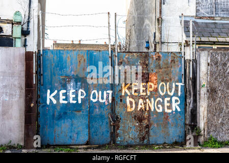 Old decaying building with warnings on gate to keep out. Danger keep out rough painted notice on rusting gates with barbed wire. Urban decay Stock Photo