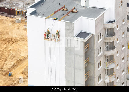 workers in a crane paint the wall of a residential high-rise building from a bird's eye view with forest on background Stock Photo