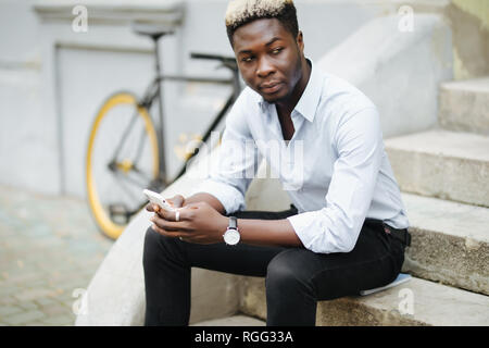 Handsome Afro American man in casual wear is using smartphone while sitting on stairs outdoors Stock Photo