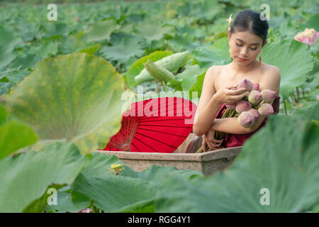 Beautiful asia women wearing traditional Thai dress and sitting on wooden boat with red paper umbrella in flower lotus lake. Her hands are holding a p
