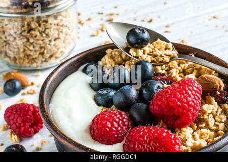 bowl of oat granola with yogurt, fresh raspberries, blueberries and nuts with cereals and spoon in a jar for healthy breakfast. close up Stock Photo