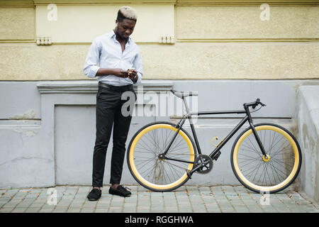 Photo of young african guy walking with bicycle outdoors on the street using mobile phone. Stock Photo