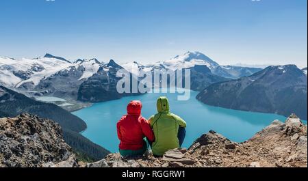 View from Panorama Ridge trail, Two hikers sitting on a rock with Garibaldi Lake, turquoise glacial lake, Guard Mountain and Stock Photo