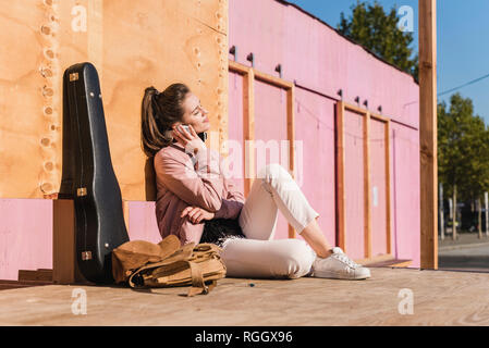Smiling young woman sitting on platform next to guitar case listening to music Stock Photo