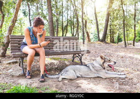 Young woman sitting on a bench in the woods looking at cell phone while her dog lying on the ground Stock Photo