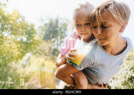 Mother carrying daughter piggyback in garden drinking a smoothie Stock Photo