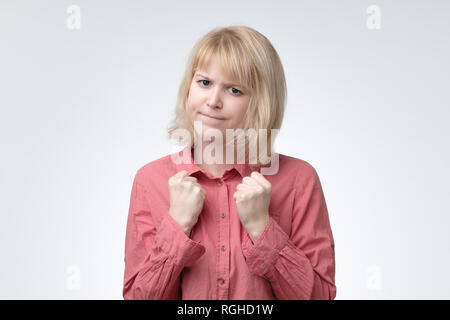 Shy woman with blonde hair and serious look keeping fists clenched in front of her defending herself Stock Photo