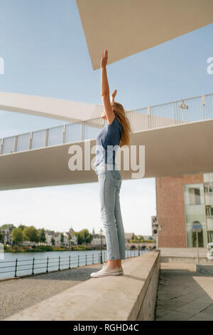 Netherlands, Maastricht, young woman standing on a wall at the riverside with raised arms Stock Photo