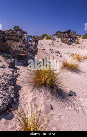 White sand dunes with rocks and bright orange grass at the coastline of Crete Island. Sea is visible through the hole in rock. Clear blue sky. Vertica Stock Photo