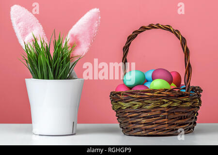 Sticking rabbit ears of a flower pot with artificial green grass, next to a basket with colored dyed eggs. Easter concept. Stock Photo