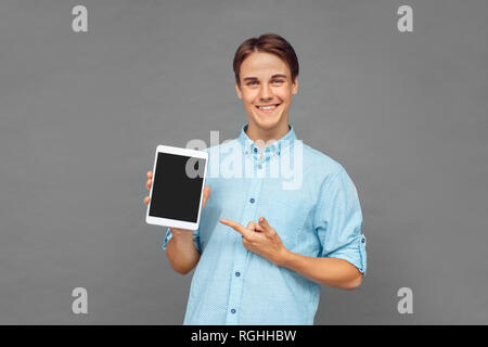 Young man standing isolated on grey wall holding digital tablet pointing at screen looking camera smiling cheerful Stock Photo
