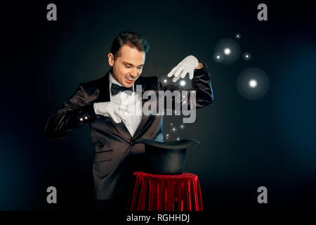 Professional magician wearing suit and gloves standing isolated on black and red background making trick with top hat on table smiling playful Stock Photo