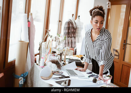 Creative person at design studio. Young african woman, near work table with different craft items . She standing near dress mannequin Stock Photo
