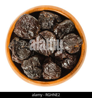 Prunes, dried plums in wooden bowl. Uncooked, dehydrated, pitted fruits of Prunus domestica with black color, used as snack. Isolated macro photo. Stock Photo