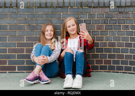 Portrait of two smiling girls sitting in front of brick wall taking selfie with smartphone Stock Photo