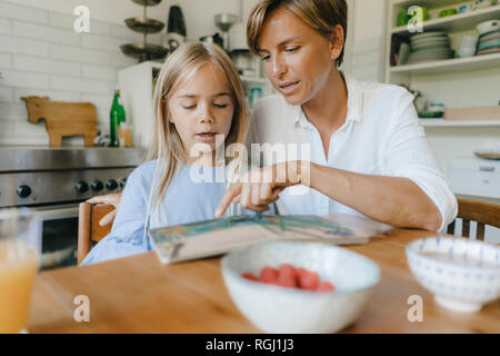Mother and daughter reading book at table at home together Stock Photo