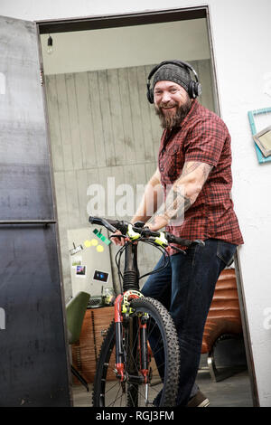 Portrait of happy man with bicycle wearing headphones in office Stock Photo