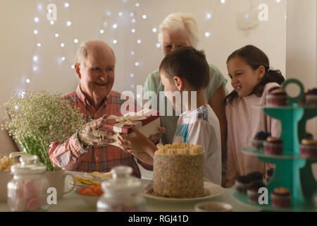 Happy multi-generation family giving present to birthday boy at home in front of a table with sweet food Stock Photo