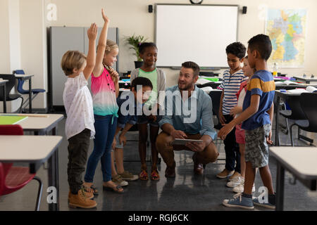 Front view of school kids raising hands while teacher teaching in classroom of elementary school Stock Photo