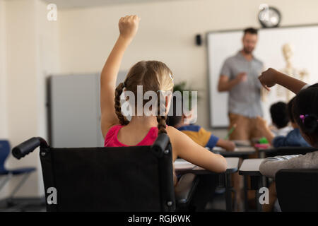 Rear view of disable schoolgirl raising hand in classroom Stock Photo