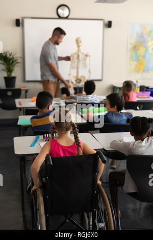 Rear view of disable schoolgirl with classmate studying in classroom sitting at desk of elementary school Stock Photo