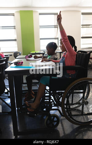 Side view of disable schoolgirl raising hand and sitting at desk in classroom Stock Photo