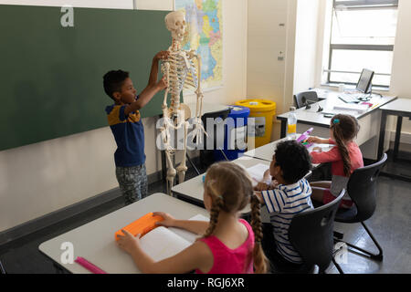 Side view of schoolboy explaining human skeleton model in classroom Stock Photo