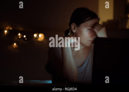 Tired woman working overtime on laptop computer at night in her home office, face is illuminated by the blue light of the screen Stock Photo