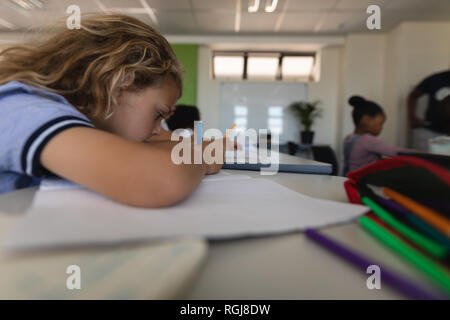 Side view of schoolgirl leaning on desk and writing on notebook in classroom Stock Photo